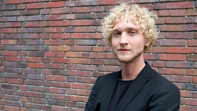 Person mit lockigem blondem Haar in schwarzem Blazer steht vor einer Ziegelwand.
